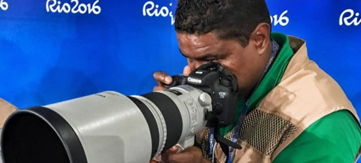 João Maia trabalhando no Rio2016 sentado de costas para um painel azul olhando no visor da câmera profissional com uma lente fixa de 300 mm da série branca da Canon . João usa camiseta verde e colete de imprensa marrom.