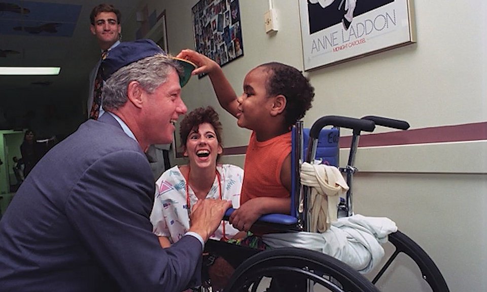 Φωτογραφία του Προέδρου Ουίλιαμ Τζ. Κλίντον να χαιρετά ασθενείς στην Παιδιατρική Πτέρυγα του Πανεπιστημιακού Νοσοκομείου Robert Wood Johnson στο New Brunswick του Νιου Τζέρσεϊ. Ένα μικρό κορίτσι παίζει με το καπέλο τουΠροέδρου Κλίντον.