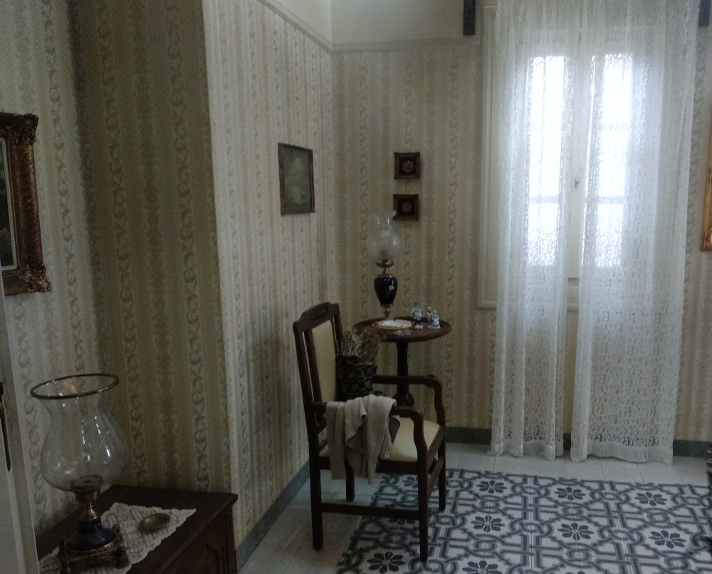 Μια ξύλινη καρέκλα πλάι στο στρογγυλό τραπεζάκι με την περίτεχνη λάμπα στο σαλόνι της οικογένειας Σεβαστού
