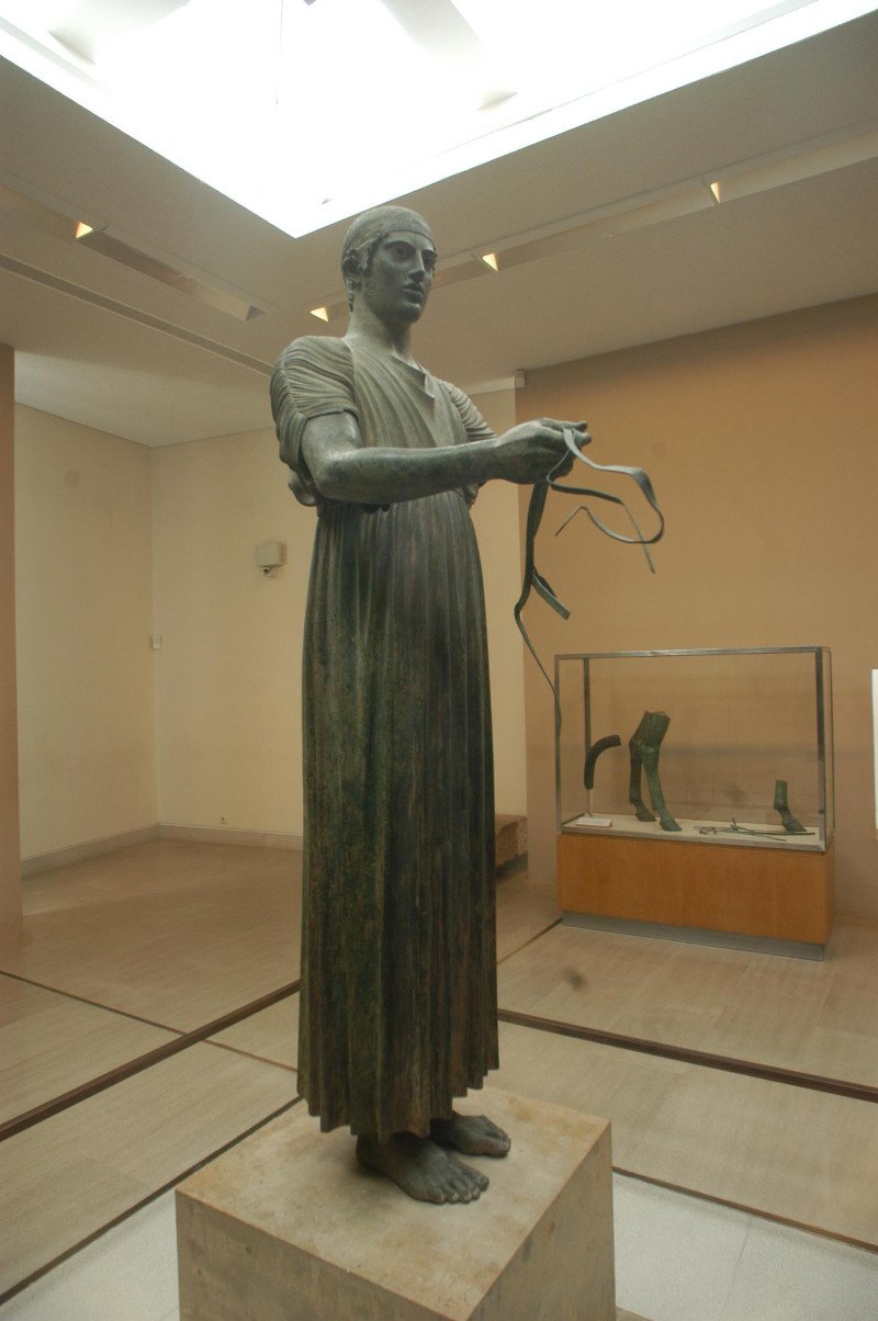 Ο Ηνίοχος οράει τον τυπικό μακρύ χιτώνα, ένας πλατύς ζωστήρας σφίγγει το χιτώνα ψηλά πάνω από τη μέση, και ένας ιμάντας σταυρώνει στο πίσω μέρος, κάτω από τις μασχάλες