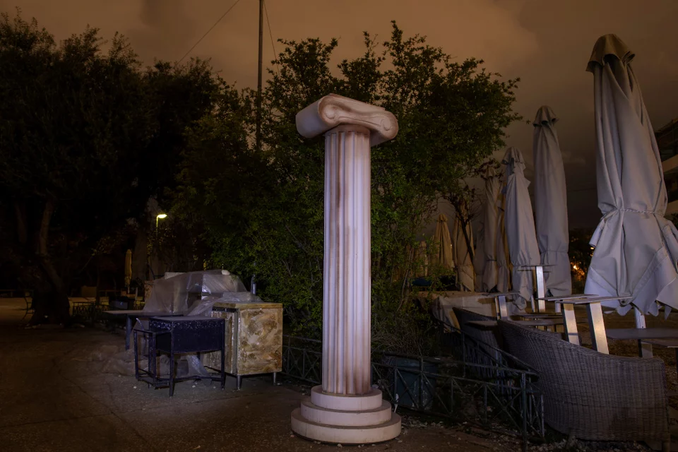 Ένα αντίγραφο μιας αρχαίας ελληνικής στήλης στέκεται δίπλα σε αποθηκευμένα τραπέζια και διπλωμένες ομπρέλες στις 18 Μαρτίου 2021, έξω από ένα κλειστό κατάστημα στην Αθήνα, όπου εστιατόρια, μπαρ και καφετέριες είχαν κλείσει από τον Νοέμβριο του 2020 για να περιοριστεί η εξάπλωση του COVID-19. Φωτογραφία: AP Photo/Petros Giannakouris