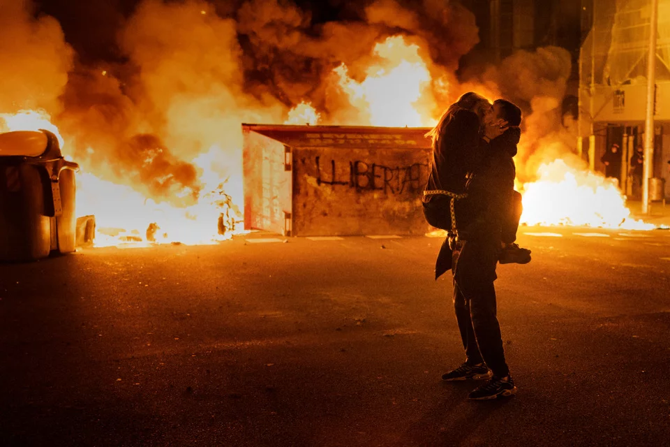 Ένα ζευγάρι φιλιέται μπροστά από ένα οδόφραγμα που πυρπολήθηκε από διαδηλωτές, κατά τη διάρκεια συγκρούσεων με την αστυνομία, μετά από διαμαρτυρία για την καταδίκη της φυλάκισης του τραγουδιστή της ραπ Πάμπλο Χασέλ, στη Βαρκελώνη της Ισπανίας, στις 18 Φεβρουαρίου 2021. Ο Χασέλ είχε καταδικαστεί για προσβολή της ισπανικής μοναρχίας και εξύμνηση της τρομοκρατικής βίας. Φωτογραφία: AP Photo/Emilio Morenatti 