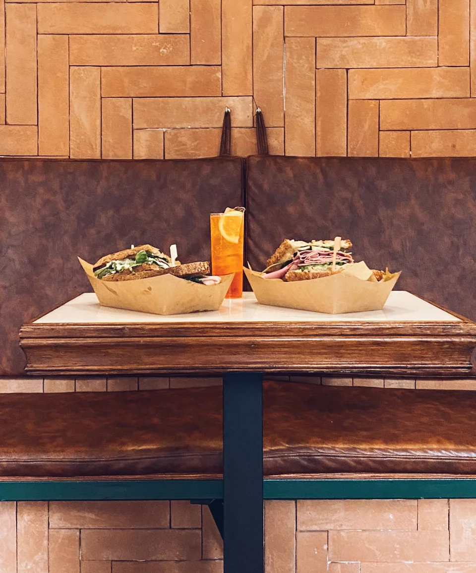 Εκτός από τα τραπεζάκια στο πεζοδρόμιο, έξω, σε μια εσοχή του τοίχου, υπάρχει και ένας καναπές με δερμάτινα μαξιλάρια, όπου μπορείς να απολαύσεις ένα Aperol Spritz τρώγοντας το σάντουιτς που επέλεξες και χαζεύοντας τους περαστικούς.