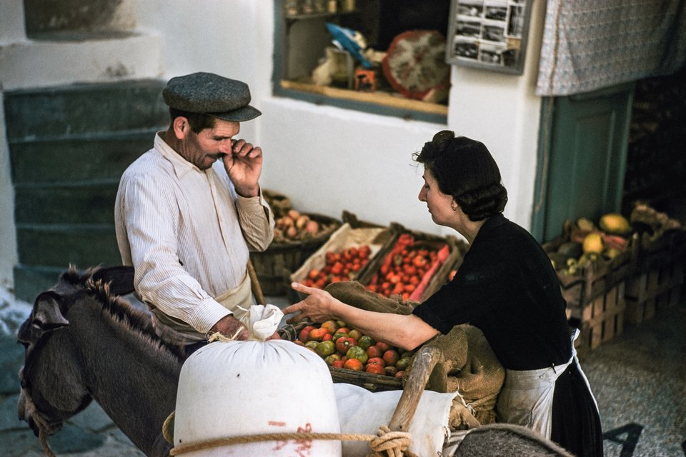 Μύκονος, 1957. Μια σοβαρή διαπραγμάτευση βρίσκεται σε εξέλιξη μεταξύ ενός αγρότη που έφερε προϊόντα στην αγορά στη Χώρα με το γαϊδουράκι του και της καταστηματάρχη Ασημίνας Κουσαθανά. Το μικρό ράφι με τις καρτ ποστάλ υποδηλώνει έναν κόσμο που αλλάζει