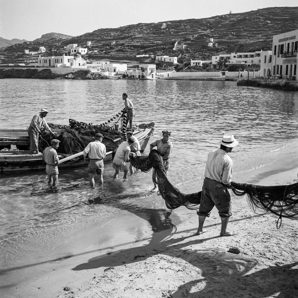 Μύκονος, 1955. Ξημερώματα στον μόλο Καμπάνη, ψαράδες ξεφορτώνουν τα δίχτυα τους για στέγνωμα και επισκευή. Το νεόκτιστο ξενοδοχείο «Λητώ» διαφαίνεται στο βάθος. Το «Λητώ» μαζί με το «Αμφιτρύων» στο Ναύπλιο ήταν τα πρώτα δύο ξενοδοχεία χτισμένα από τον ΕΟΤ το 1952-53.