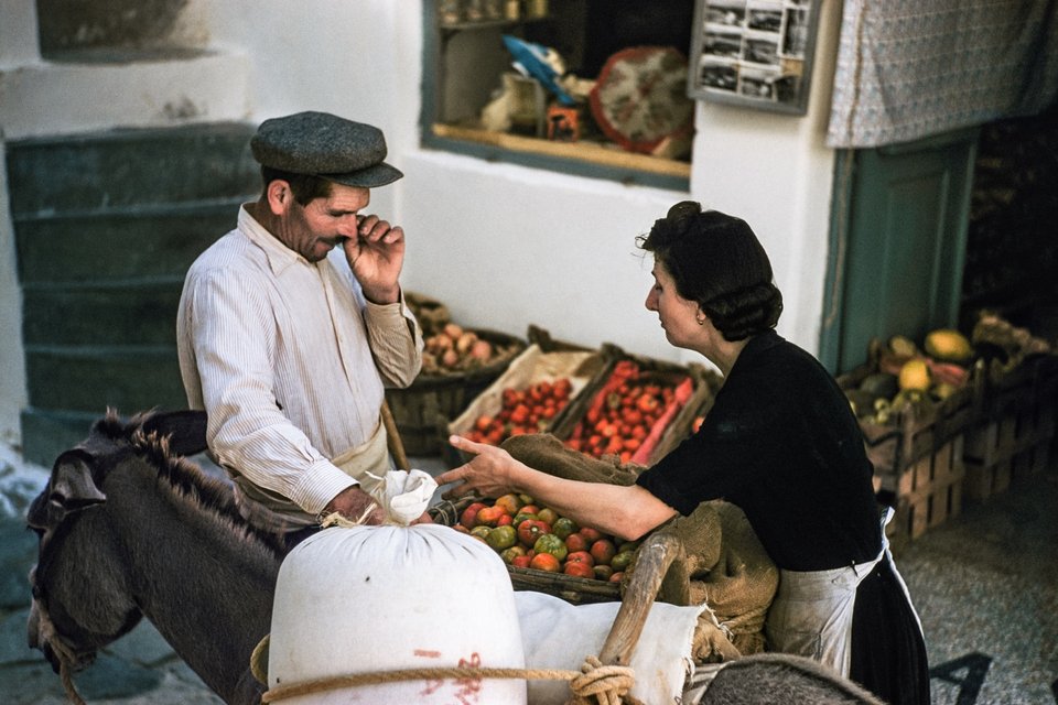 Μύκονος, 1957. Μια σοβαρή διαπραγμάτευση βρίσκεται σε εξέλιξη μεταξύ ενός αγρότη που έφερε προϊόντα στην αγορά στη Χώρα με το γαϊδουράκι του και της καταστηματάρχη Ασημίνας Κουσαθανά. Το μικρό ράφι με τις καρτ ποστάλ υποδηλώνει έναν κόσμο που αλλάζει.