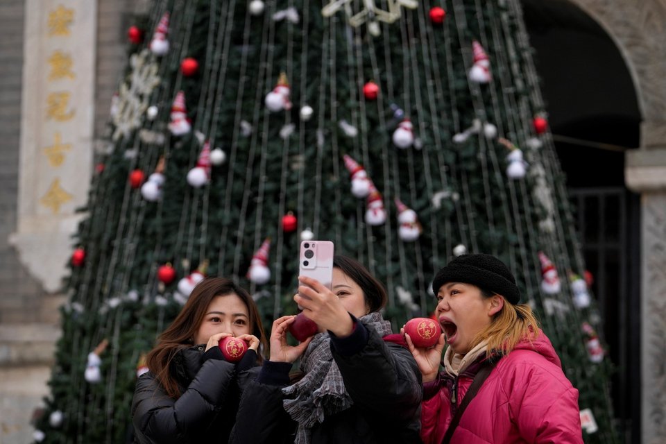 Γυναίκες κρατούν μήλα με τους κινεζικούς χαρακτήρες καθώς βγάζουν selfie μπροστά από ένα χριστουγεννιάτικο δέντρο στο Πεκίνο / AP Photo/Andy Wong