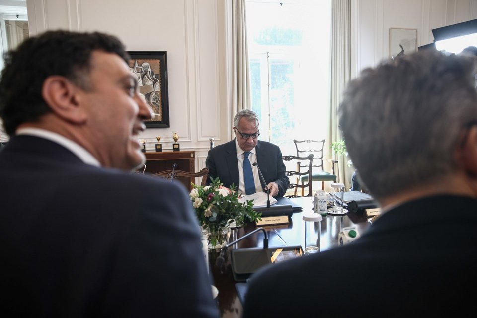 Ο Μάκης Βορίδης κοιτάζει τα χαρτιά του, ενώ απέναντί του υπουργοί της κυβέρνησης δείχνουν να συζητούν σε εύθυμο κλίμα