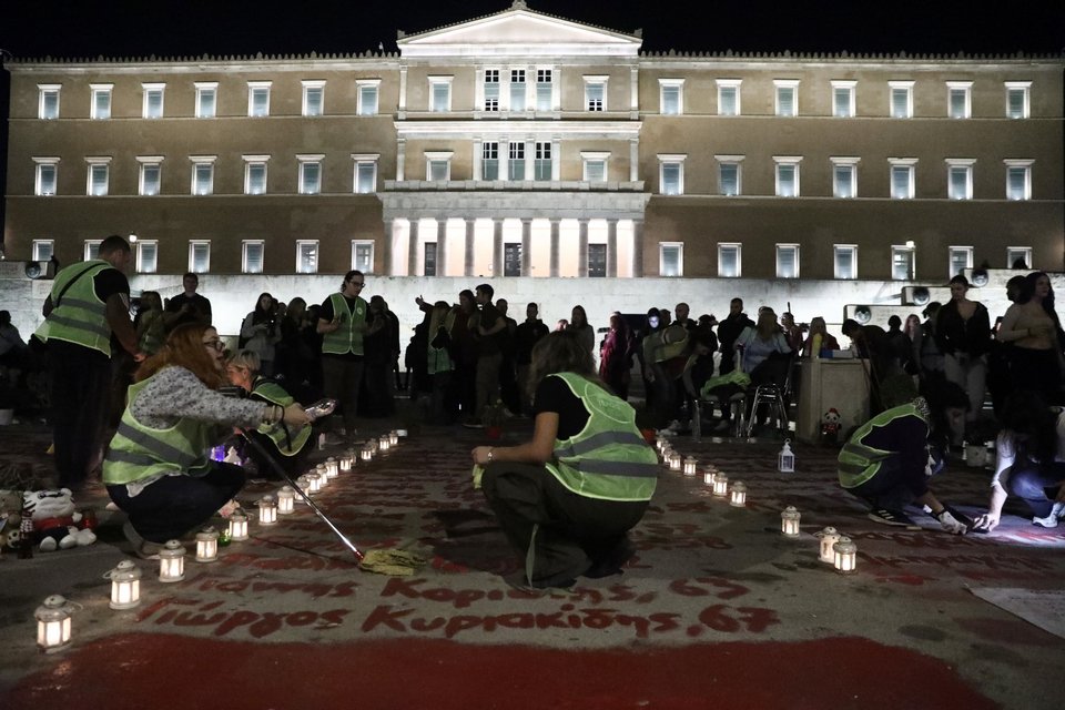 Πολίτες με κίτρινα γιλέκα καθαρίζουν τον χώρο όπου είναι γραμμένα τα ονόματα των 57 θυμάτων των Τεμπών / Intime
