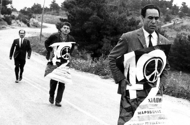 Ο Γρηγόρης Λαμπράκης συνεχίζει την Πορεία, τυλιγμένος με την αφίσα της Ειρήνης. Τον ακολουθούν οι Ανδρέας Μαμωνάς και Μπάμπης Παπαδόπουλος