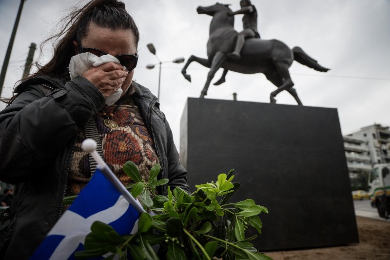 Μια γυναίκα κλαίει, κρατώντας μαντήλι στο ένα χέρι, λουλούδια και σημαία στο άλλο