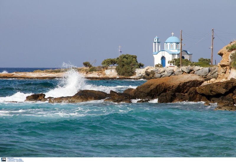 Κύματα σκάνε στα βράχια της Ικαρίας με φόντο ένα εκκλησάκι πλάι στη θάλασσα