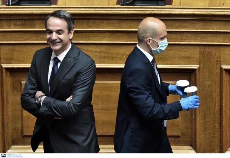 Ο Κυριάκος Μητσοτάκης χαμογελά ενώ υπάλληλος της Βουλής με μάσκα καθαρίζει το βήμα 