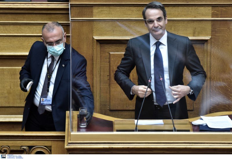 Ο Κυριάκος Μητσοτάκης ετοιμάζεται να  πάρει τον λόγο, ενώ υπάλληλος της Βουλής με μάσκα καθαρίζει το βήμα 