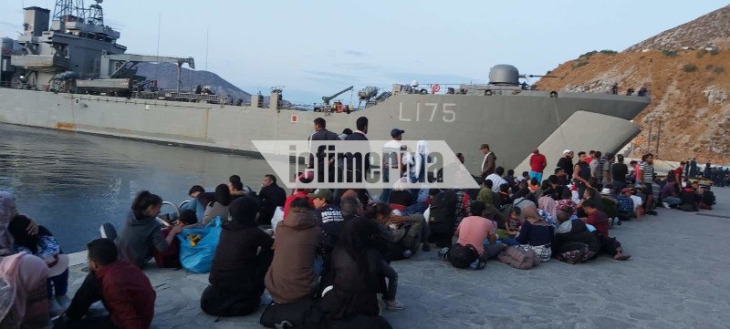 Πρόσφυγες καθιστοί μπροστά από πλοίο του Πολεμικού Ναυτικού