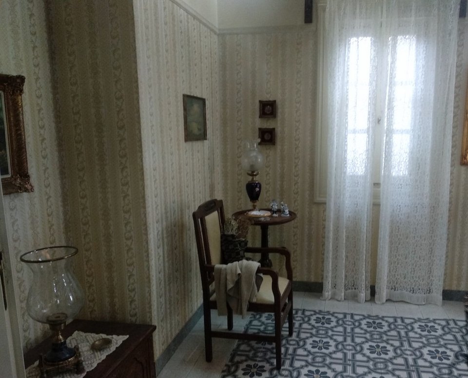 Μια ξύλινη καρέκλα πλάι στο στρογγυλό τραπεζάκι με την περίτεχνη λάμπα στο σαλόνι της οικογένειας Σεβαστού