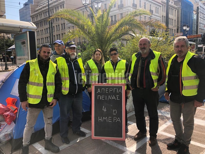 Οι συμβασιούχοι στέκονται στο πεζοδρόμιο μπροστά από ταμπέλα που γράφει: Απεργία πείνας για 4η ημέρα
