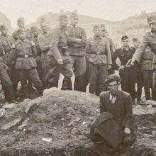 Ένας άνδρας γονατιστός στο χείλος ενός λάκκου γεμάτου πτώματα, κι ένας Γερμανός στρατιώτης με το χέρι σηκωμένο, το πιστόλι να ακουμπά σχεδόν τον σβέρκο του / WIKIPEDIA