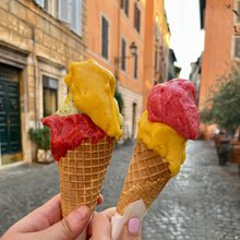 Δύο άτομα κρατούν πολύχρωμα παγωτά gelato σε ένα λιθόστρωτο δρόμο στη Ρώμη, με ιστορικά κτίρια στο παρασκήνιο