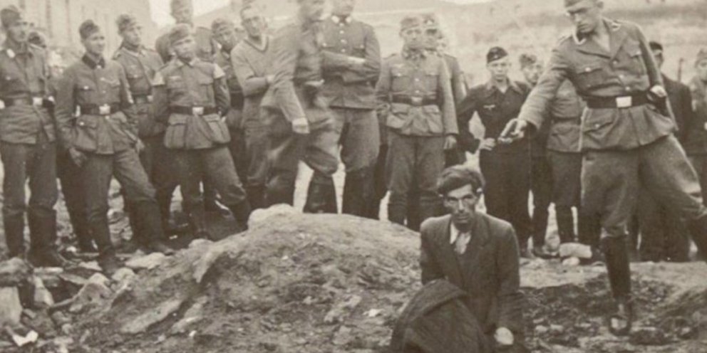 Ένας άνδρας γονατιστός στο χείλος ενός λάκκου γεμάτου πτώματα, κι ένας Γερμανός στρατιώτης με το χέρι σηκωμένο, το πιστόλι να ακουμπά σχεδόν τον σβέρκο του / WIKIPEDIA