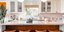 Κουζίνα διαμερίσματος με λευκά ντουλάπια, πάγκο, φυτό, φωτιστικά, καρέκλες