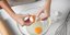 Γυναίκα σπάει αυγά σε διάφανο μπολ σε λευκό τραπέζι 