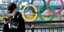 Νεαρός με μάσκα κοιτάζει το κινητό του με φόντο τους Ολυμπιακούς κύκλους