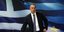 Ο Χρήστος Σταϊκούρας στέκεται όρθιος μπροστά από την ελληνική σημαία