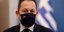 Στέλιος Πέτσας με μάσκα και σακάκι με ελληνική σημαία πίσω