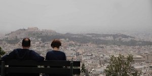 Ενα ζευγάρι κάθεται σε παγκάκι στον λόφο του Λυκαβηττού και ατενίζει την Αθήνα, που έχει καλυφθεί από την αφρικανική σκόνη