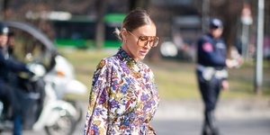 Γυναίκα διασχίζει το δρόμο και φοράει φλοράλ φόρεμα με λευκές μπότες και λευκή τσάντα