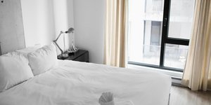 Ενα κρεβάτι με λευκά σεντόνια και παράθυρο με μπεζ κουρτίνες
