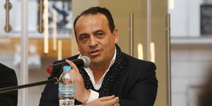 Ο Βασίλης Ζούλιας με μαύρο σακάκι κάθεται και μιλά στο μικρόφωνο