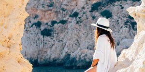 Γυναίκα ντυμένη στα λευκά κάθεται σε ένα βράχο φορώντας καπέλο