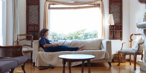 Mια γυναίκα κάθεται στον καναπέ του σπιτιού, μπροστά από το παράθυρο