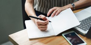 Γυναίκα σημειώνει σε τετράδιο δίπλα στο κινητό της και τον υπολογιστή