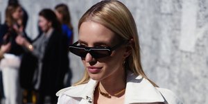 Mια γυναίκα με μαύρα γυαλιά, λευκό τοπ, παντελόνα και ελαφριά καπαρντίνα