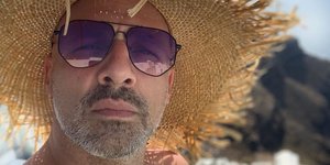 Ο Νίκος Μουτσινάς με ψάθινο καπέλο και φόντο την Σαντορίνη