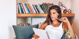 Η Χριστίνα Μπόμπα κάθεται σε λευκή πολυθρόνα με μια βιβιλιοθήκη από πίσω της