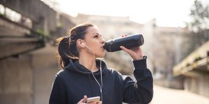 Γυναίκα με μαύρο φούτερ πίνει νερό από μπουκάλι και κρατάει το κινητό της