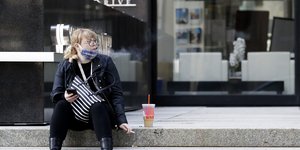 Γυναίκα με μάσκα και καφέ, κάνει τσιγάρο καθήμενη σε πεζούλι