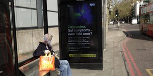 Ηλικιωμένη σε στάση του Λονδίνου, μπροστά από αφίσα του NHS που προειδοποιεί για τις συνέπειες του κορωνοϊού