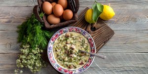 Πιάτο με μαγειρίτσα σε ξύλο κοπής μπροστά από καλάθι με αυγά, λεμόνια και μυρωδικά