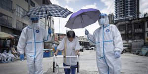 Εργαζόμενοι στη Μαλαισία με στολές και μάσκα, κρατούν ομπρέλες πάνω από ηλικιωμένη που μόλις υποβλήθηκε σε τεστ για τον κορωνοϊό