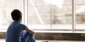 Παιδί καθισμένο στο πάτωμα πλάι σε λούτρινο κουκλάκι κοιτά έξω από το παράθυρο