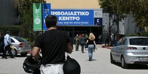 άνδρας με κράνος μηχανής στο χέρι κοντά σε αυτοκίνητο με κόσμο γύρω του έξω από εμβολιαστικό κέντρο στο Περιστέρι