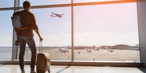 άνδρας με βαλίτσα κοντά σε τζάμι σε αεροδρόμιο με αεροπλάνο να πετάει