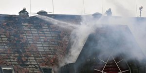 Πυροσβέστες σε στέγη οικήματος προσπαθούν να σβήσουν φωτιά ρίχνοντας νερό με μάνικες