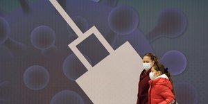 γυναίκες με μάσκες περπατούν μπροστά από εικόνα μιας τεράστιας σύριγγας