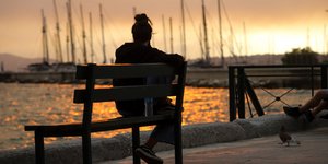 Κοπέλα κάθεται σε παγκάκι και απολαμβάνει το ηλιοβασίλεμα στη θάλασσα